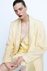 Vestido corto color amarillo pálido, de satén con tirantes finos, escote en V y bajo asimétrico con abertura. Presenta detalles de encaje en el escote, lateral y ruedo.