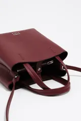 Bolso pequeño tipo tote o cubo de piel vacuna texturizada color negro, con asas cortas superiores y correa larga ajustable para llevar al hombro o cruzado. Presenta el logo metálico de Bimba y Lola en dorado en el frente.