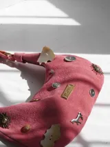 Cartera baguette de gamuza rosa con asa corta y múltiples apliques decorativos: botones, monedas, dijes de metal con forma de sol y caballo, y placa metálica con la marca "Agnes Lenoble".