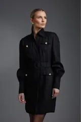 Vestido camisero corto negro con cuello, cierre frontal con botones dorados, cinturón con hebilla circular y mangas largas abullonadas con puños abotonados. Presenta bolsillos con solapa en el pecho y en la falda.