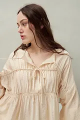 Blusa color beige de caída fluida con cuello redondo y lazos frontales. Diseño con cortes horizontales y frunces, mangas largas con puños elastizados.