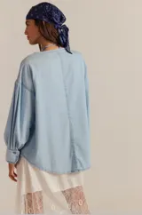 Camisa de denim chambray celeste, estilo oversized, con escote henley alargado con botones, hombros caídos y mangas abullonadas con puños terminados en volados.
