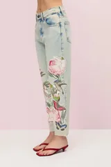 Pantalón de jean celeste con lavado vintage y calce mom. Presenta parches bordados con motivos florales, de mariposas y colibríes. El ruedo está desflecado.