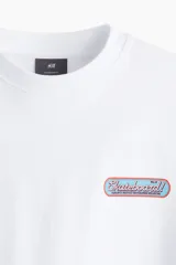 Remera blanca de algodón pesado con cuello redondo ribeteado, hombros caídos, bajo recto y estampado pequeño en el pecho. Corte oversized.