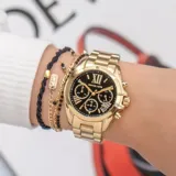 Reloj Michael Kors modelo Bradshaw, con caja y correa de acero inoxidable dorado, esfera negra con números romanos y tres subesferas. Incluye cronógrafo y fechador.