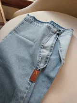 Pantalón de jean celeste con cintura elastizada, corte recto, bolsillos laterales y aplique de cuero marrón en el bolsillo delantero. Presenta botones metálicos en los laterales de la cintura y etiqueta de cuero en la parte trasera.