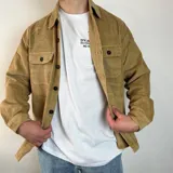 Sobrecamisa color crema de pana con cuello clásico, cierre frontal con botones negros, dos bolsillos de parche en el pecho con solapa abotonada y puños abotonados.