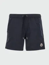Traje de baño tipo boxer de color azul marino, con cintura elástica y cordón de ajuste. Presenta un pequeño parche circular con el logo de Moncler en la parte inferior derecha del frente.
