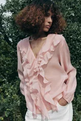 Camisa color rosa pálido confeccionada en tejido ligero con lyocell. Presenta escote en V, cierre frontal con botones, mangas largas abullonadas con puños elásticos y detalle de volantes en el frente.