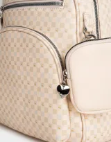 Mochila color crema con estampado geométrico de cuadros, confeccionada en cuerina. Tiene asa corta de mano, correas regulables, doble bolsillo frontal con cierre (uno con dije de corazón) y pouch lateral extraíble con cierre y dije de corazón.