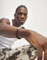 Pulsera de cuero trenzado color marrón oscuro con un cilindro central de aleación de metales bañada en plata de ley, con textura rugosa y plateada. Cierre tipo mosquetón de metal plateado.