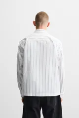 Camisa cropped fit confeccionada en tejido de algodón a rayas verticales negras sobre fondo blanco. Cuello solapa y manga larga acabada en puño con botones. Bolsillos frontales de plastrón con solapa. Cierre frontal de botonadura.