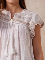 Blusa color marrón claro de algodón con bordados y calados blancos. Tiene cuello a la base, escote en V con lazo de ajuste y mangas cortas amplias.
