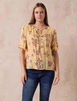 Blusa amarilla estampada con flores rosadas, de gasa, con cuello a la base, escote en V plisado y lazo de ajuste. Delantera con detalles en puntilla y mangas 3/4 con puño fruncido.