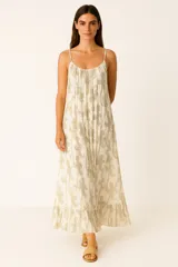 Vestido largo de viscosa con breteles finos ajustables y escote redondo. El tejido es blanco con un estampado repetitivo de figuras estilo paisley en color dorado o beige brillante (lurex). La falda es amplia y termina con un pequeño volante fruncido en el ruedo.