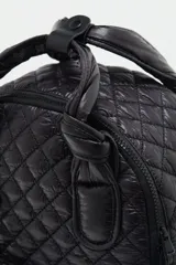 Mochila negra matelassé con correa ajustable y manijas para llevar al hombro. Tiene cierre de cremallera, accesorio colgante decorativo, bolsillo interno y espacio para laptop.