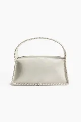 Cartera de mano color crudo, con forma rectangular y asa corta. Presenta detalle de tira entrelazada en los bordes y cierre superior.
