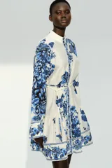 Vestido corto camisero blanco con estampado floral azul, cuello mao, botonadura frontal y cinturón fino para atar extraíble. Mangas globo de corte raglán, puños abotonados y costura fruncida bajo la cadera.