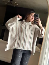 Sweater tejido color beige con cuello alto y mangas largas. Presenta aberturas verticales con detalles en marrón y negro.