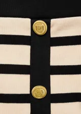 Minifalda de punto a rayas horizontales en color crema y negro, con ribetes en contraste. Presenta dos bolsillos de solapa con botón dorado en la parte delantera y botones decorativos.