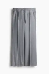 Pantalón gris de sarga con cintura elástica fruncida y cordón ajustable. Piernas amplias con pliegues cosidos, bolsillos laterales discretos y bolsillos decorativos atrás.