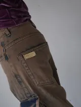 Pantalón cargo de jean color marrón con efecto desgastado, múltiples bolsillos funcionales y detalles metálicos en bronce.