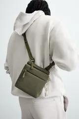 Bandolera negra con acabado engomado y textura acanalada. Tiene un compartimento principal con cierre de solapa y botón a presión, un bolsillo interior con cremallera y un monedero cuadrado en la correa. La correa de hombro es ajustable.