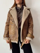 Campera marrón de gamuza sintética con interior de piel sintética color beige, cierre cruzado con cremallera, cuello con solapas amplias y cinturón con hebilla en puños y ruedo.