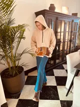 Sweater beige de corderito con capucha y bolsillo frontal de cuero en tonos marrón y ocre. Presenta tajos laterales y cierre parcial con cremallera en el cuello.