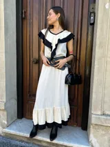 Vestido midi color crudo con detalles en negro en cuello, volados y ruedo. Incluye cinturón ancho negro con tachas y hebilla dorada.