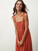 Vestido midi de color naranja, con escote recto y tirantes finos que se anudan sobre los hombros. Posee una cintura entallada con frunces que dan paso a una falda fluida.