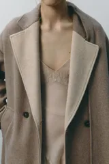 Abrigo largo color beige confeccionado en lana. Presenta cuello solapa con bufanda desmontable, manga larga, bolsillos laterales y cierre frontal cruzado con botones.