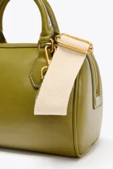 Bolso formato mini bowling color verde oliva con detalles metálicos dorados en las asas. Tiene doble asa de mano y correa bandolera extraíble. Incluye bolsillo interior con cremallera y cierre principal con cremallera.