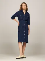 Vestido camisero midi de denim azul oscuro, con cuello clásico, manga tres cuartos y cierre frontal con botones. Incluye cinturón de la misma tela para ajustar a la cintura y dos bolsillos de parche con solapa en el pecho.