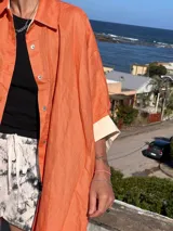 Camisa de lino color naranja durazno, de corte amplio y manga larga con puños anchos que contrastan en color crema/blanco. Presenta cuello camisero y cierre frontal con botones claros.