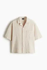 Camisa de manga corta color crema en tejido sirsaca de algodón. Modelo boxy con cuello abierto, tapeta lisa, bolsillo superior y bajo recto. Corte holgado.