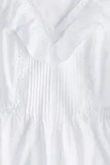 Blusa blanca sin mangas de tejido liviano de algodón con alforzas e inserciones de encaje, escote en V con ribete de volados y peplum con leve vuelo.
