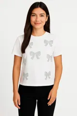 Remera blanca de algodón con cuello redondo y mangas cortas. Estampado con moños plateados brillantes.