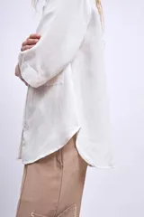 Camisa blanca de manga tres cuartos, con cuello camisero y corte holgado. Presenta un bolsillo de parche en el pecho izquierdo.