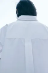 Camisa blanca oversized de popelina de algodón, con cuello clásico, botones frontales, hombros caídos, mangas largas con puños abotonados, canesú con pliegue en la espalda y bajo redondeado.