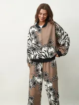 Campera bomber con estampado de flores en blanco y negro sobre fondo beige y marrón. Tiene cuello, puños y bajo acanalados en negro, y cierre frontal con cremallera.