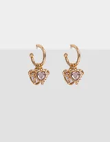 Aros dorados tipo argolla con dijes colgantes de corazón de perla, corazón de strass rosa y moño dorado.