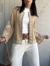 Chaqueta color beige con bordados blancos en el frente y las mangas, de corte recto y mangas anchas.