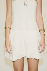 Vestido corto color crudo, de gasa, con escote en V y tirantes finos regulables. Presenta cordón cruzado con lazada en el delantero y falda con volantes. Cierre trasero con cremallera oculta.