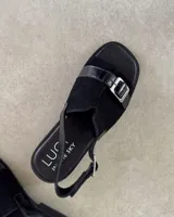 Sandalias negras con suela plana, tira ancha de gamuza con hebilla plateada y tira trasera ajustable.