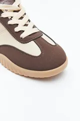 Zapatilla urbana de mujer con silueta retro, combinando paneles de gamuza marrón oscuro y material color crema en el cuerpo. Presenta detalles en color bordó en el talón y cordones beige. La suela es robusta de color marrón claro con relieve.
