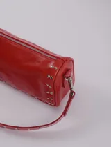 Bandolera color rojo de cuero con tachas plateadas. Tiene dos asas desmontables, una corta con tachas para llevarla en la mano o al hombro y una larga para llevarla al hombro o cruzada. Tiene bolsillo interior con cierre y forro.