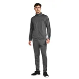 Conjunto deportivo Under Armour de hombre, color gris, compuesto por campera con cierre y pantalón largo. La campera tiene cuello alto, cierre frontal completo, bolsillos laterales y logo de la marca en el pecho. El pantalón tiene cintura elástica con cordón ajustable y logo de la marca en la pierna.