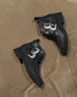 Botín corto negro de cuero vacuno, estilo cowboy o biker, con hebillas plateadas decorativas en la caña y tachas plateadas en la puntera. Posee un taco bajo y cuadrado.