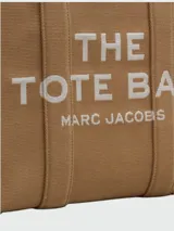 Bolso tote grande de tela negra con asas de mano y hombro. Presenta el texto "THE TOTE BAG MARC JACOBS" en color blanco en el frente. Los bordes laterales tienen una cinta blanca con texto repetido.